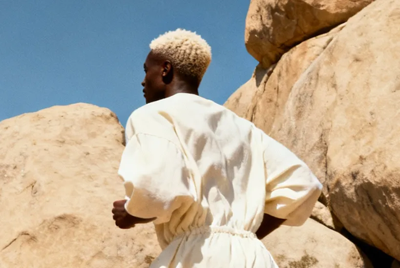 Person with short blonde hair wearing a white dress running outdoors near large rocks under a clear blue sky.
