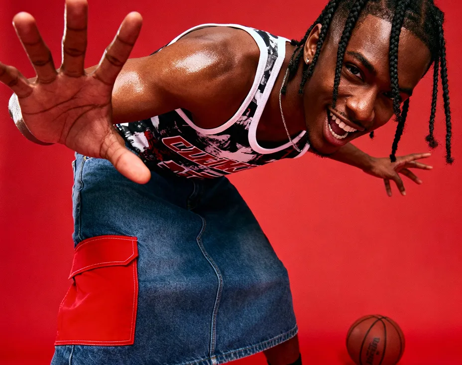 Smiling man with braided hair in a sleeveless shirt and denim skirt playing basketball against a red background.