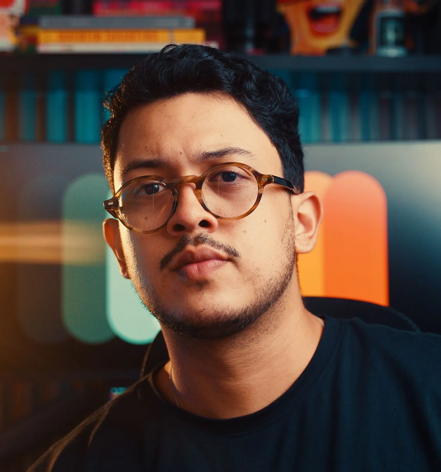 Portrait of a man with short curly hair and glasses, wearing a dark shirt, with a colorful blurred background.