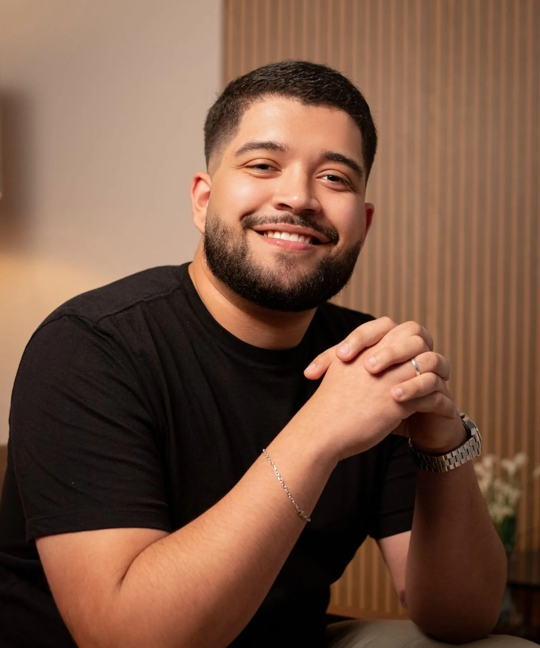 Smiling man with short black hair and beard wearing a black t-shirt, silver bracelet, watch, and ring, sitting indoors.