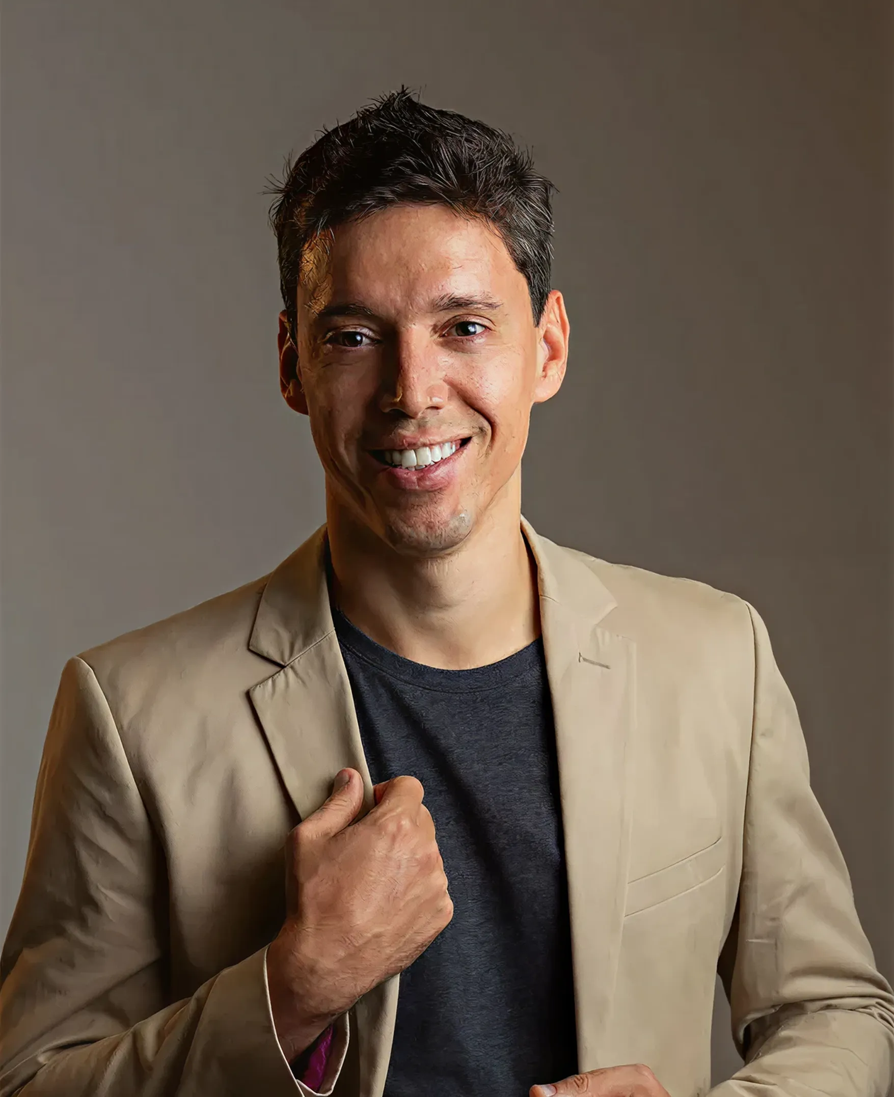 Smiling man wearing a beige blazer over a dark t-shirt against a plain background.