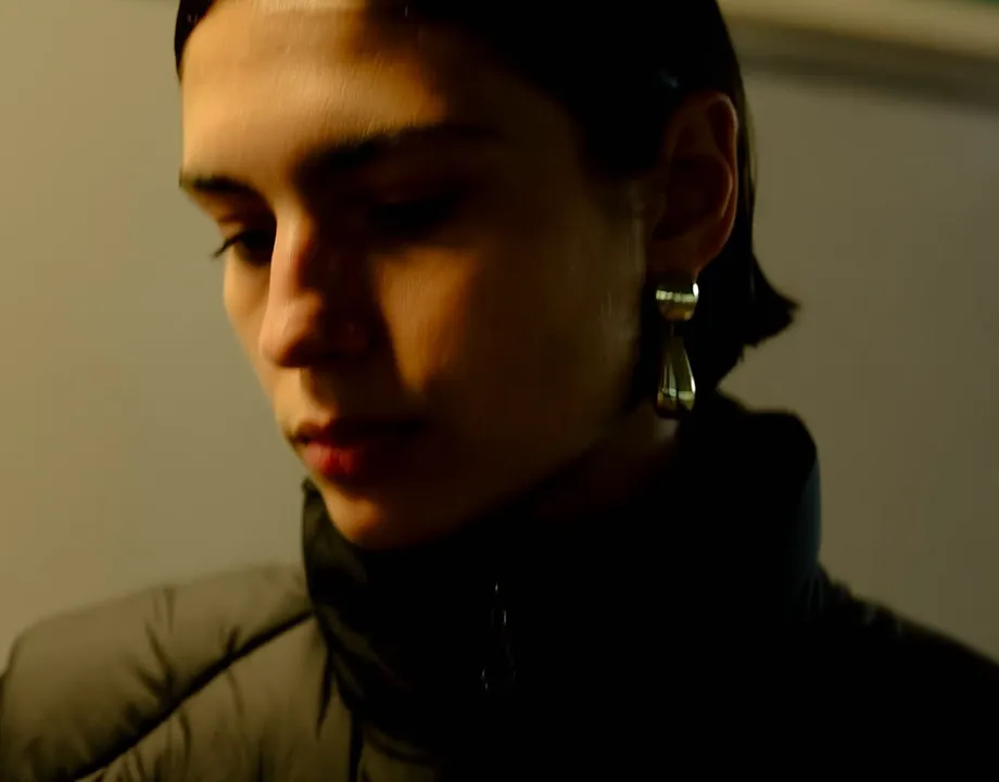 Close-up portrait of a person with short dark hair wearing silver earrings and a black jacket, looking downward in soft warm lighting.