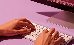 Two hands typing on a white keyboard in front of a computer monitor on a purple surface.