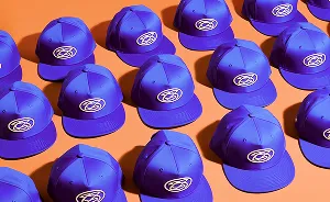 Multiple blue baseball caps with a white circular logo arranged in rows on an orange surface.