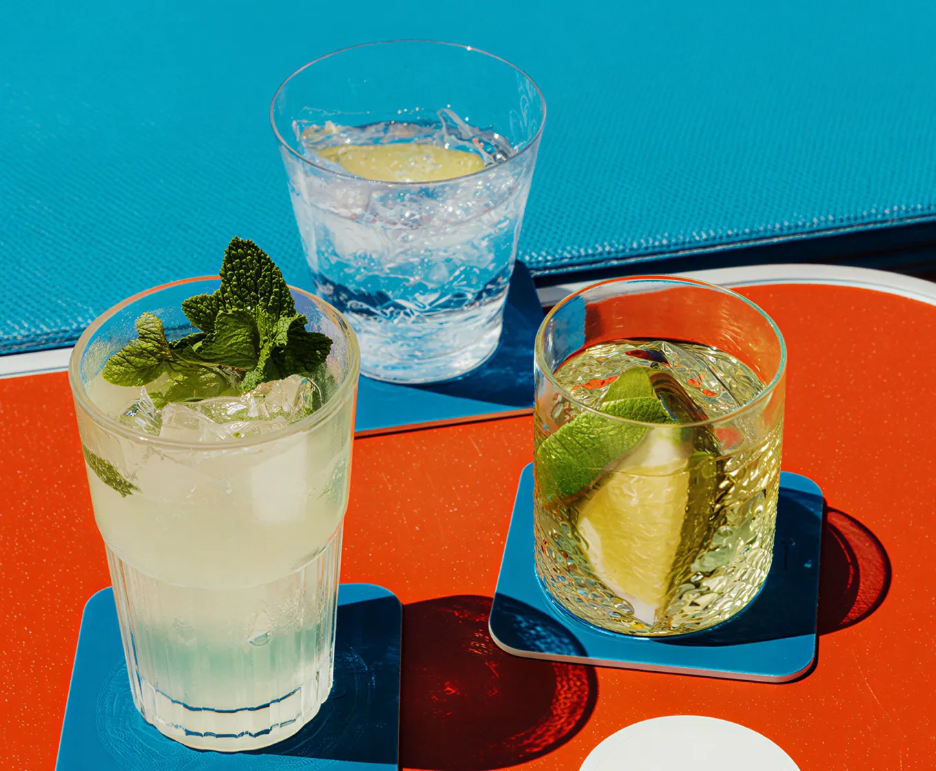 Three refreshing beverages on blue coasters on a red table, including a mint garnished drink, a clear drink with lemon slice, and a drink with lime wedge.